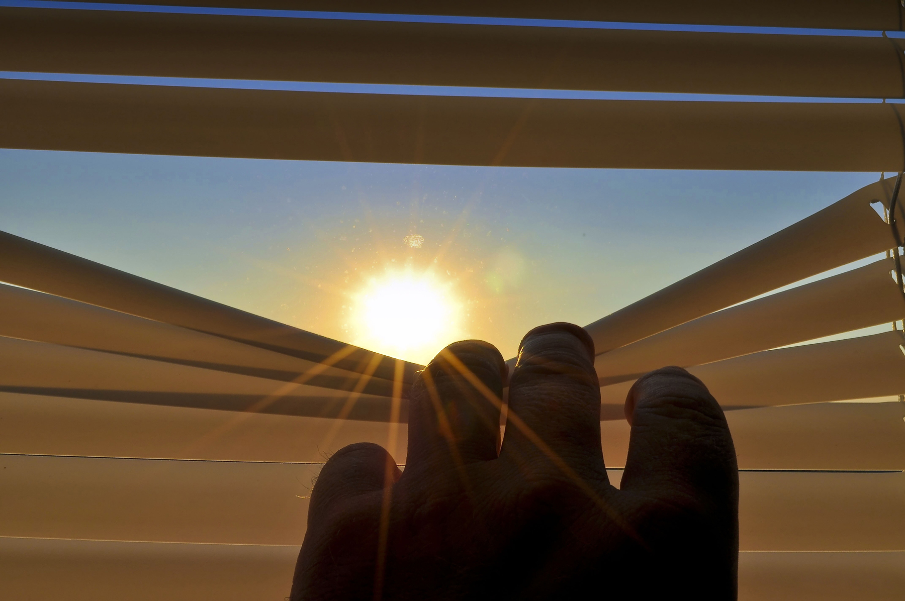 A person pulling down part of a window blind with the sun shining through